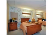 A health and rehabilitation center room with two wooden beds, each with a peach-colored bedspread. The room has floral wallpaper, a ceiling light, and wooden nightstands beside each bed. There are some decorative items on the nightstands.
