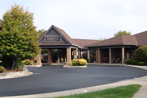Exterior view of a single-story brick building with a covered entrance and a sign that reads 'Emeritus'. The building is surrounded by trees and shrubs, with a paved driveway in front.