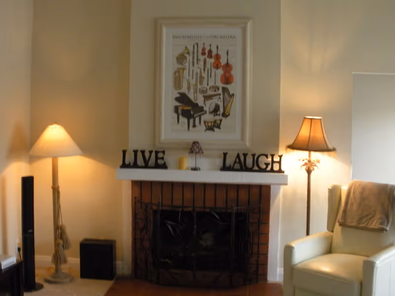 Cozy living room area with a brick fireplace in the center. On the mantel above the fireplace are decorative letters spelling 'LIVE' and 'LAUGH' with a small candle and lamp in between. A framed picture of various musical instruments hangs above the mantel. To the left of the fireplace is a tall floor lamp and a speaker. To the right is a cream-colored armchair with a blanket draped over the back and another floor lamp beside it.