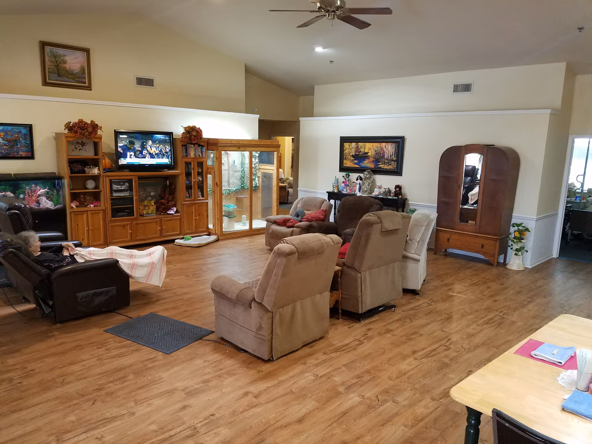 A spacious common living area with multiple recliners arranged around a TV and wooden entertainment cabinets on hardwood floors.