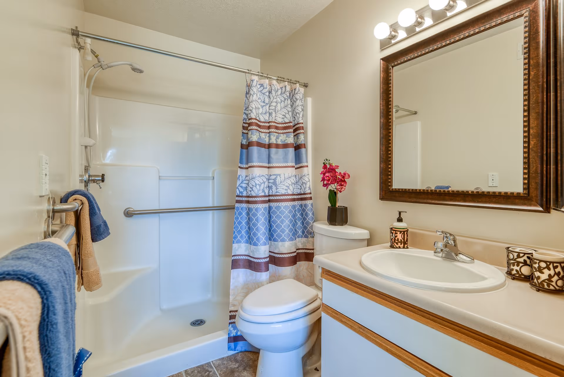 A clean bathroom featuring a white toilet, a sink with a countertop, a large framed mirror above the sink, and a shower area with a blue and brown patterned shower curtain. There are towels hanging on a metal bar on the left wall and a small potted plant with pink flowers on the toilet tank.