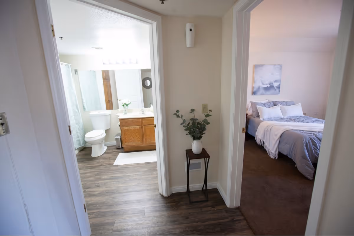 View of a hallway with two open doors, one leading to a bathroom with a toilet, sink, mirror, and shower curtain, and the other leading to a bedroom with a bed, pillows, and a wall painting. A small table with a vase and green plant is placed in the hallway between the doors.