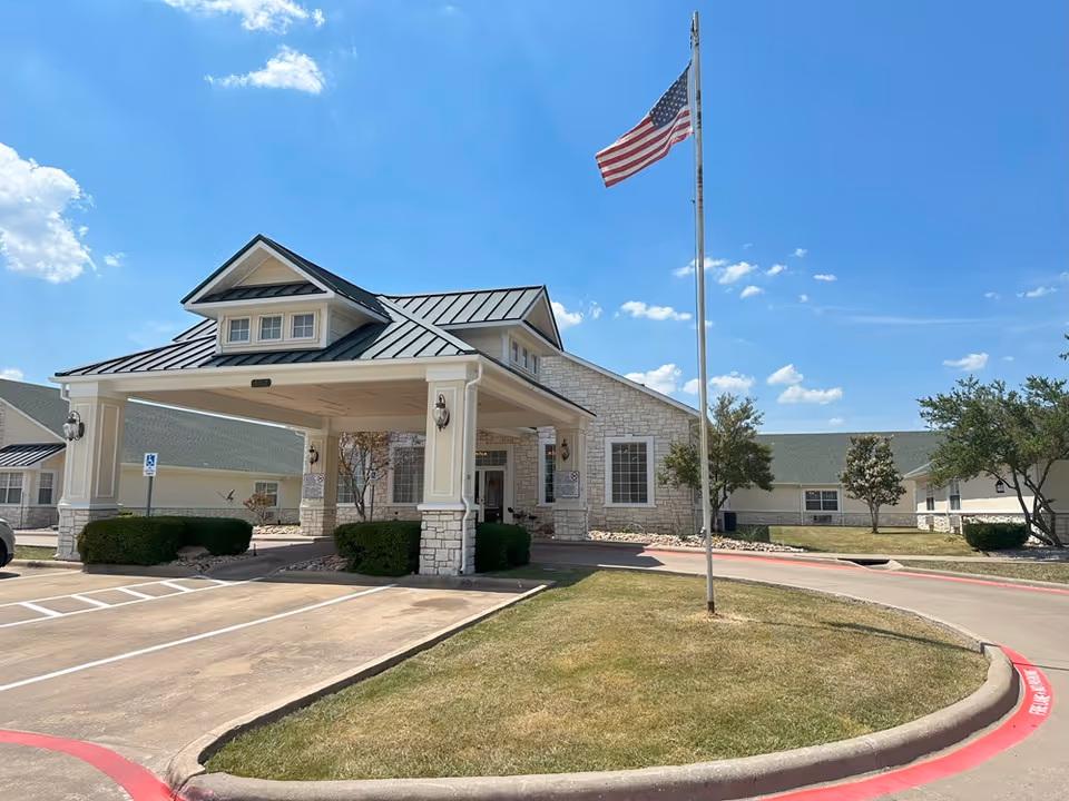 Exterior view of a senior living facility with entrance canopy, flagpole, and landscaping.