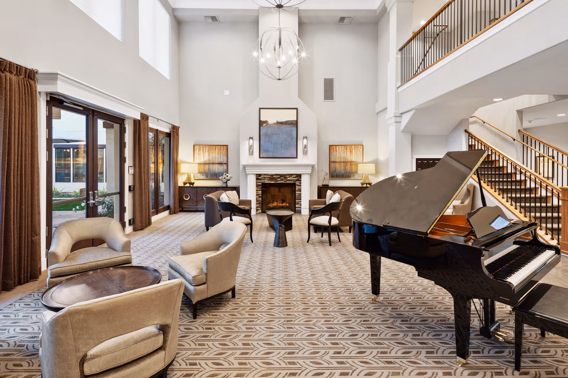 Spacious and elegant living room area with a grand piano on the right, several beige armchairs arranged around small tables, a fireplace with artwork above it, and large windows with brown curtains letting in natural light. A staircase with wooden railings is visible on the right side.