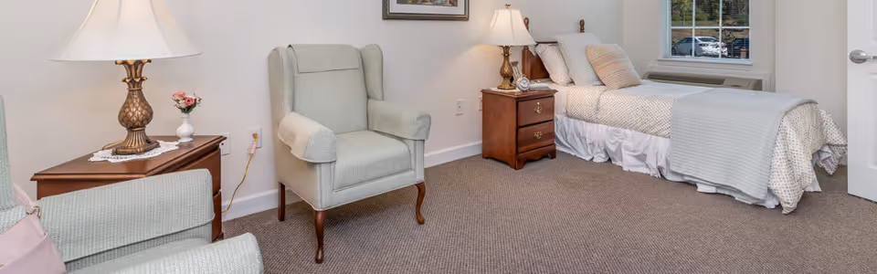 A senior living bedroom with a single bed covered in light-colored bedding, a wooden nightstand with a lamp and clock, a comfortable armchair, and a small wooden table with a lamp and flower vase. There is a window and a door visible in the room.