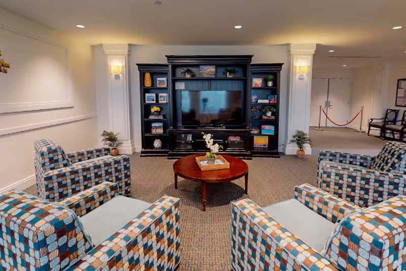 A cozy living room area with four patterned armchairs arranged around a round wooden coffee table with a small plant centerpiece. Against the wall is a large dark wood entertainment center with a flat-screen TV and decorative items on the shelves. The room has neutral-colored walls and carpet, with soft lighting and some plants near the corners.