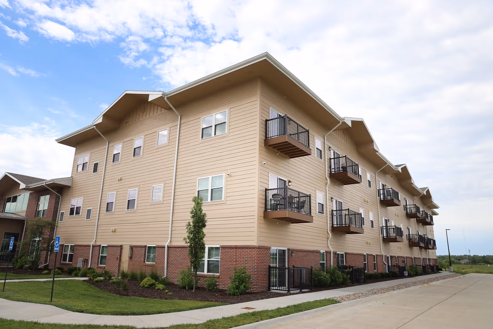 Exterior view of a three-story residential building with beige siding and brick accents, featuring multiple balconies with black railings. The building is surrounded by landscaped greenery and a paved driveway under a partly cloudy sky.