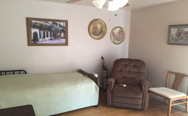 A simple bedroom with a single bed covered in a light green blanket, a brown recliner chair, and a wooden chair with a white cushion. The walls are decorated with four framed pictures, and there is a ceiling fan with lights above.