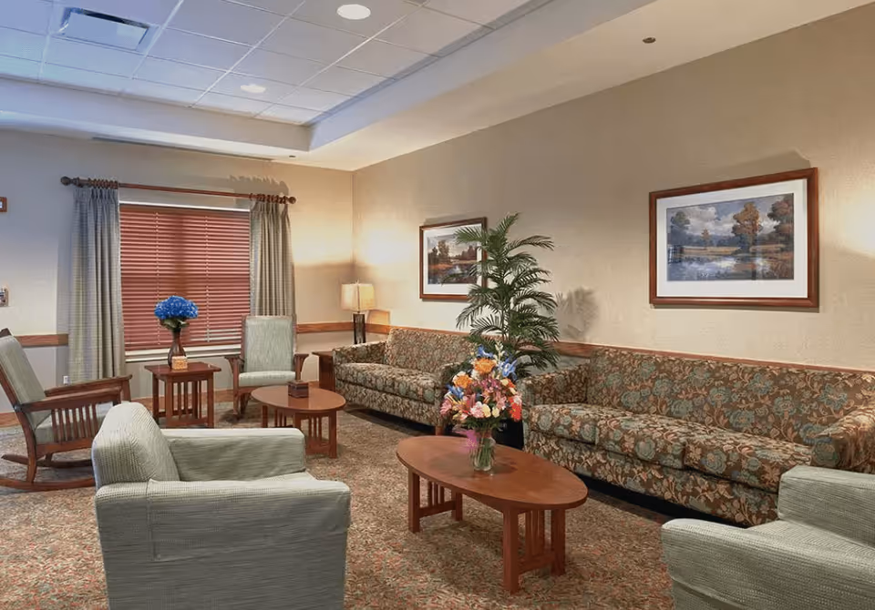 A cozy living room area with patterned sofas, two armchairs, wooden coffee tables with flower vases, a potted plant, framed landscape paintings on the wall, a window with curtains and blinds, and a floor lamp providing warm lighting.