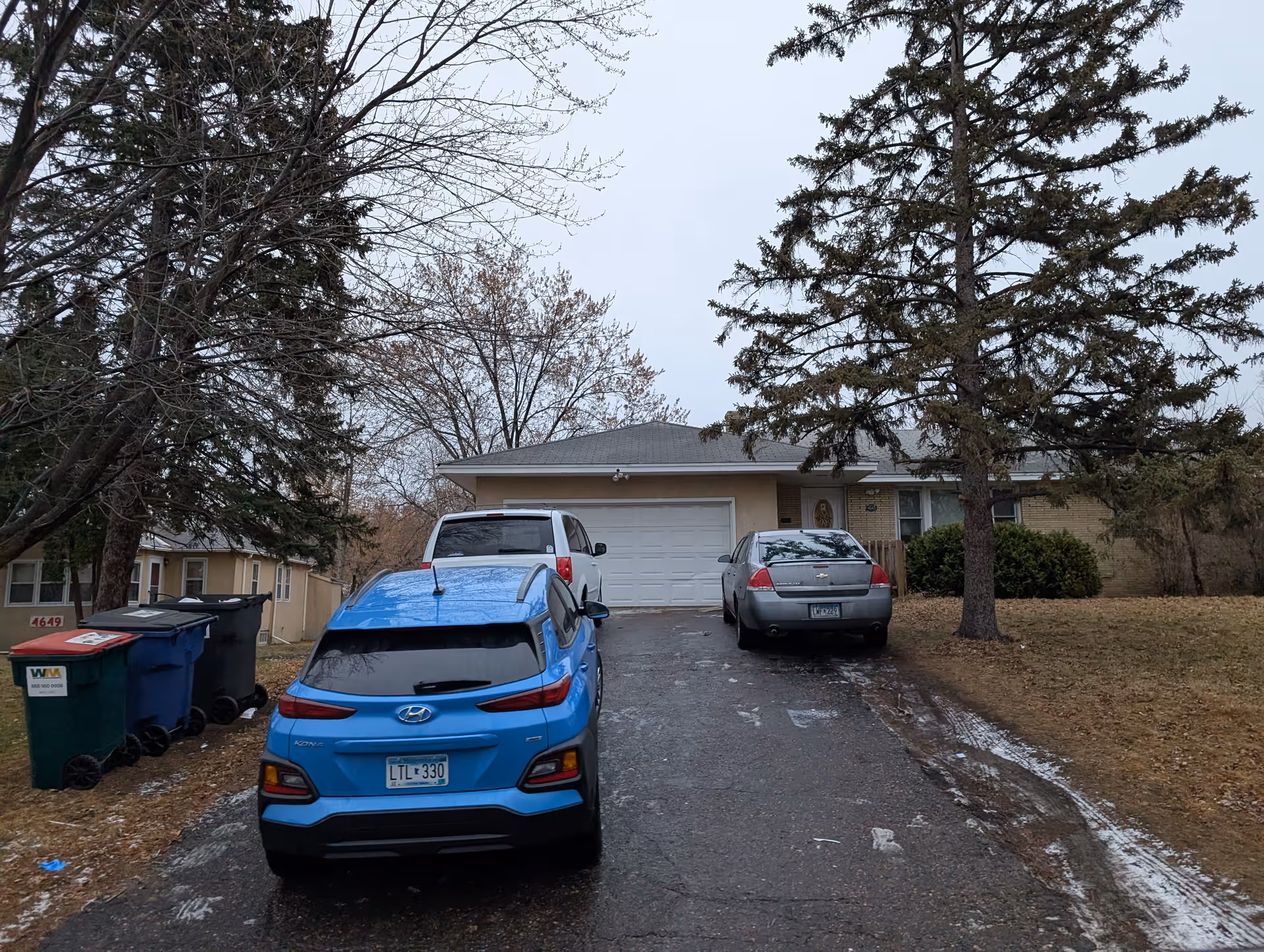 A driveway leading to a single-story house with a closed garage door. Three cars are parked on the driveway: a blue Hyundai Kona, a white van, and a gray sedan. There are several large trees around the house and some trash bins on the left side near the street. The sky is overcast and the ground appears wet with patches of ice or snow.