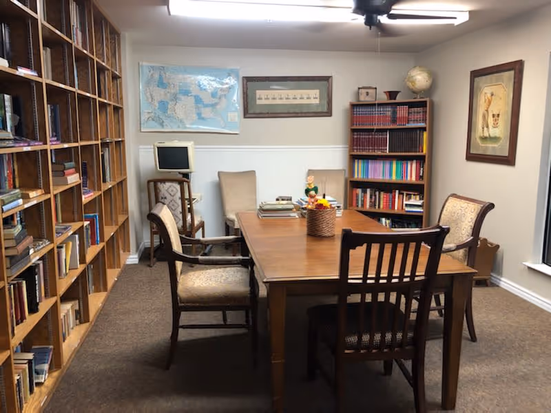A cozy library or study room with wooden bookshelves filled with books along the left wall. A wooden table with four chairs is centered in the room, with a basket of flowers and some books on the table. The back wall has a map, framed pictures, and a small computer on a stand. The room is carpeted and has a ceiling fan with lights.