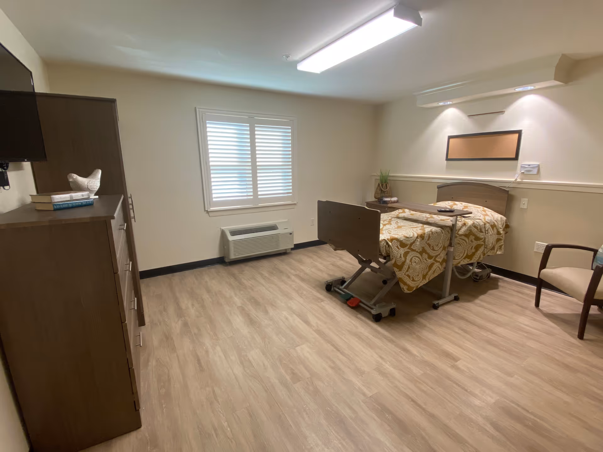 A simple, clean bedroom in a senior living facility with a single bed covered in a patterned yellow and white blanket. The room has light-colored wood flooring, a window with white shutters, a wooden dresser with books and a decorative bird on top, a wall-mounted TV, a small side table with a plant, and a chair with a cushion.