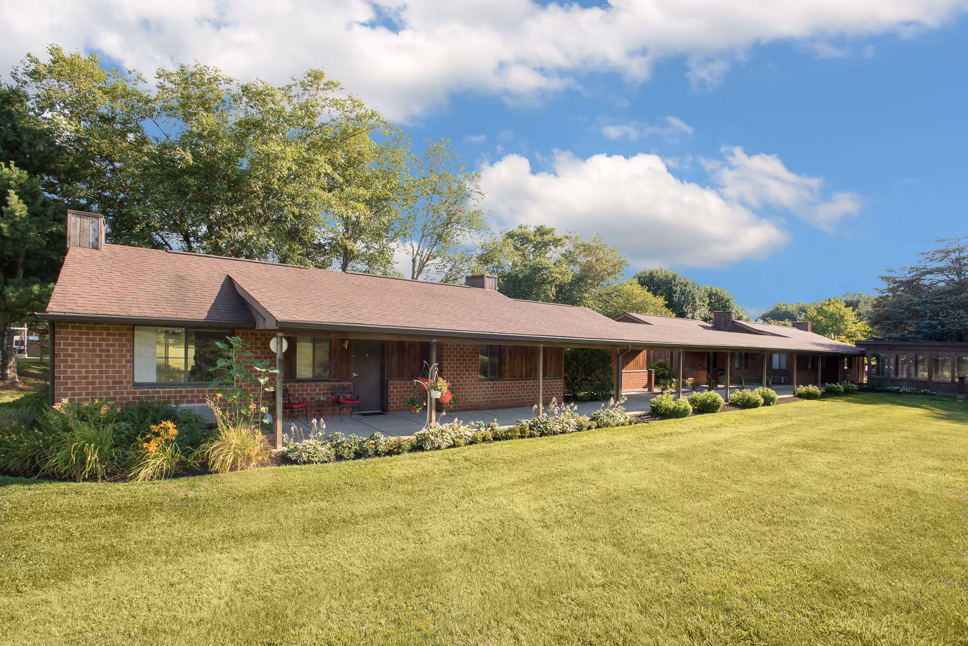 Single-story brick retirement community building with a covered walkway, landscaped flowerbeds and a large lawn under a blue sky.