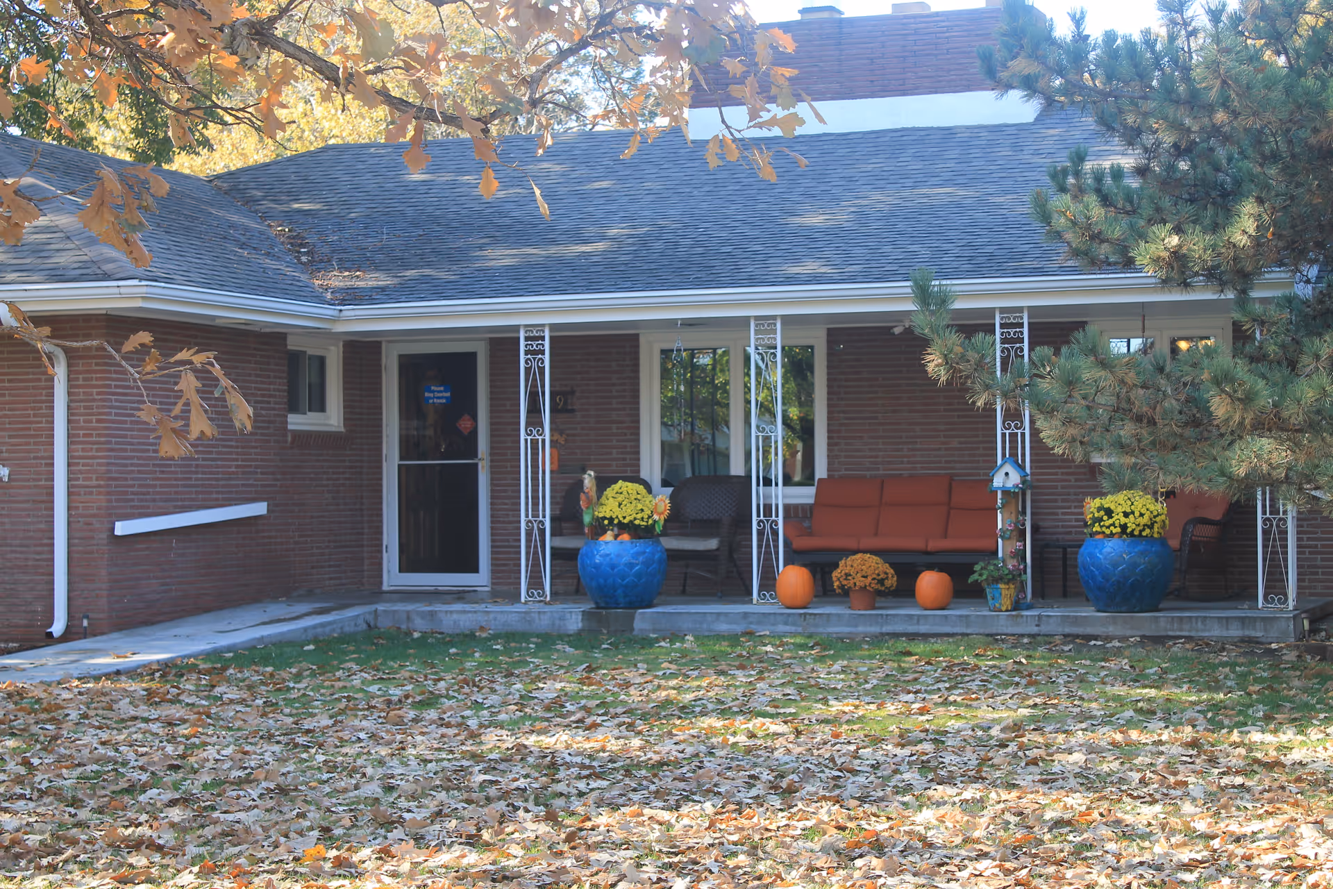 Front porch of a brick building with a covered seating area featuring orange cushioned chairs, blue flower pots with yellow flowers, pumpkins, and autumn leaves scattered on the lawn in front.