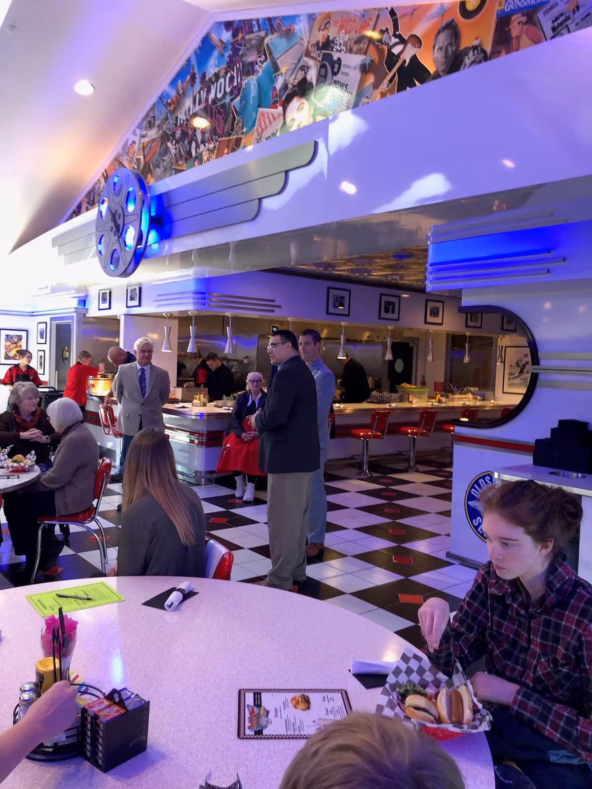 Interior of a retro-style diner with black and white checkered floor and red chairs. Several people are seated at tables eating, while others stand near the counter area. The diner features a large decorative film reel with blue lights and a colorful mural above the counter. The atmosphere is lively and nostalgic.