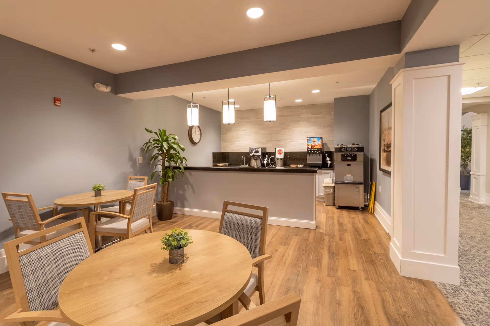 A cozy dining area with round wooden tables and cushioned chairs, a small potted plant on each table, a counter with a coffee machine and beverage dispensers, pendant lights hanging from the ceiling, a wall clock, and a potted plant in the corner. The room has wood flooring and light gray walls.