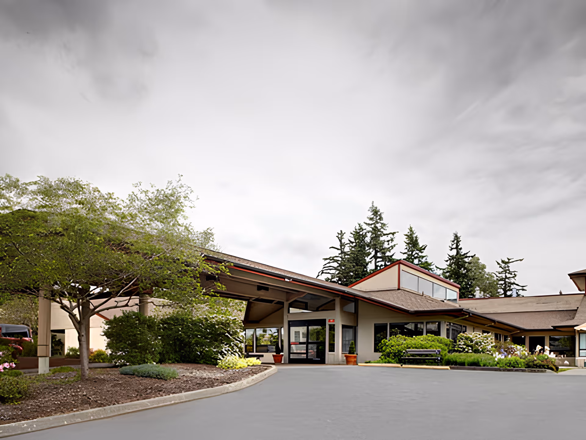 Exterior view of Avalon Healthcare - Bellingham facility showing the entrance with a covered driveway, surrounded by landscaped greenery including trees and bushes under a cloudy sky.