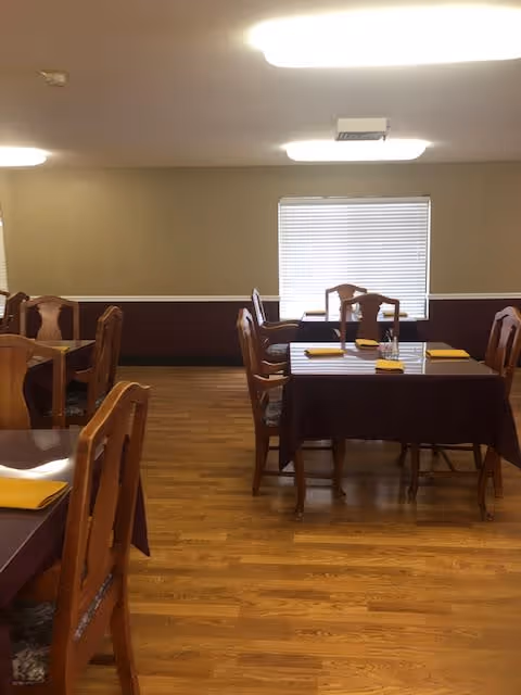 Interior view of a dining room with wooden floors and several tables covered with dark tablecloths. Each table has wooden chairs and yellow napkins placed on top. A window with closed blinds is visible on the far wall, and ceiling lights illuminate the room.