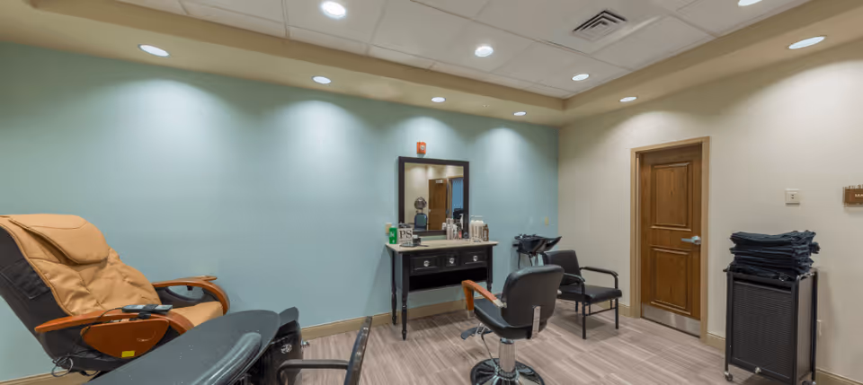 A salon-style interior room with styling chairs, a mirror and vanity, a massage chair, and stacked towels.