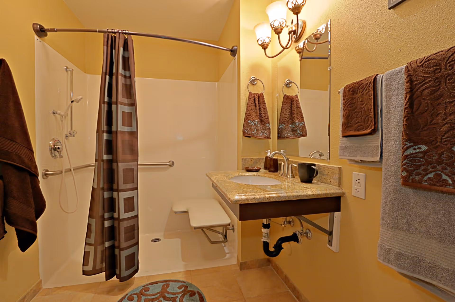 A bathroom with a walk-in shower featuring a fold-down seat and a patterned shower curtain. The room has beige walls and tiled floor. There is a granite countertop with a sink, a mirror above it, and wall-mounted light fixtures. Several brown and gray towels hang on towel rings and bars.