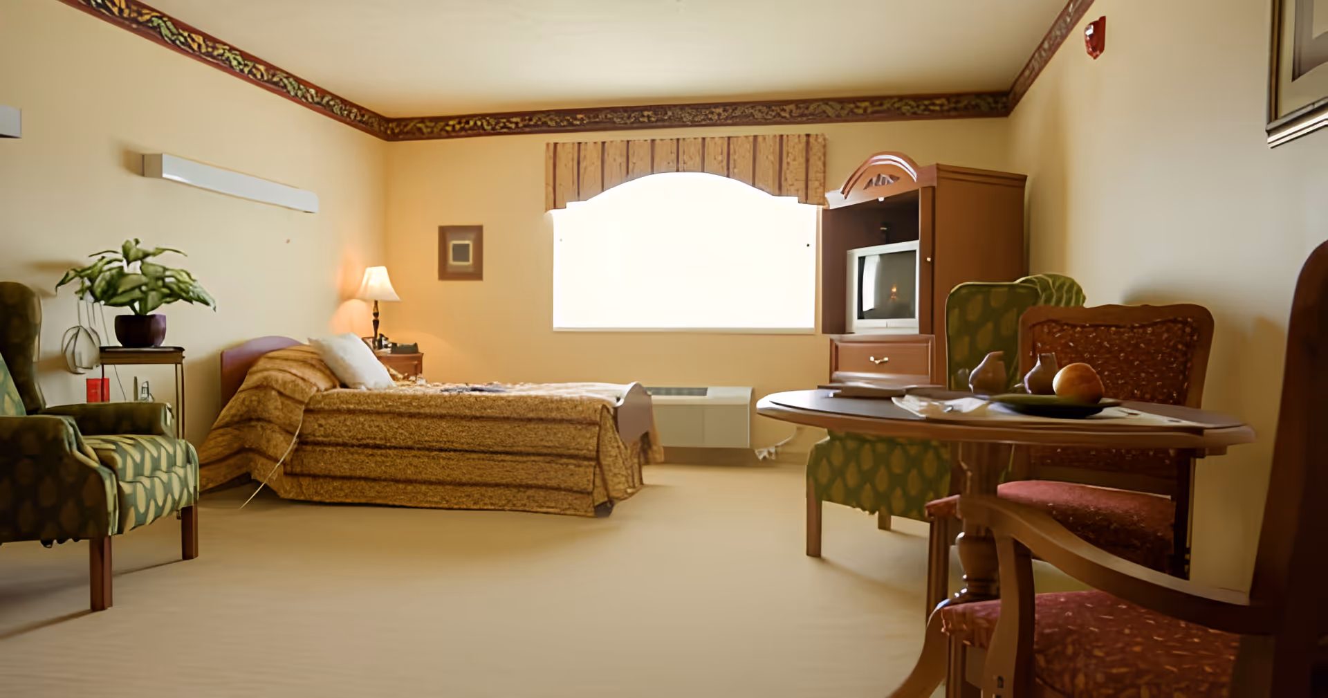 A cozy bedroom in a senior living facility with a single bed covered in a brown patterned bedspread, a bedside table with a lamp, a green armchair with a plant on a small table next to it, a wooden cabinet with a TV, and a round wooden table with chairs. The room has beige walls with decorative trim near the ceiling and a large window with a valance.
