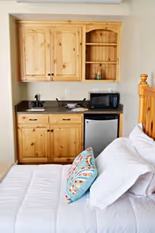 A small kitchenette area with wooden cabinets, a black countertop, a sink, a microwave, and a mini refrigerator next to a bed with white bedding and a colorful decorative pillow.