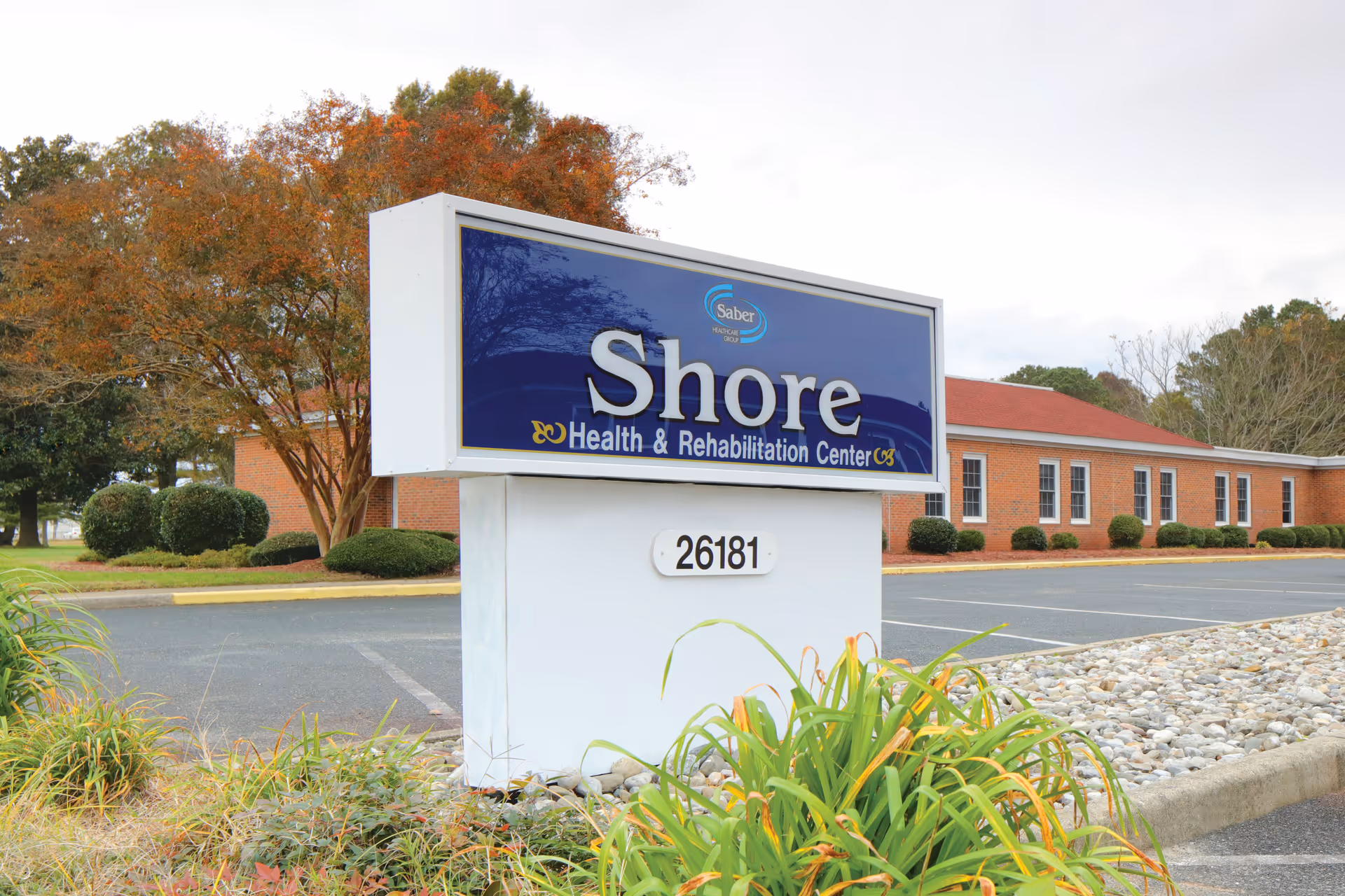 Outdoor view of the Shore Health & Rehabilitation Center sign with the building and parking lot in the background, surrounded by landscaping and trees with autumn foliage.