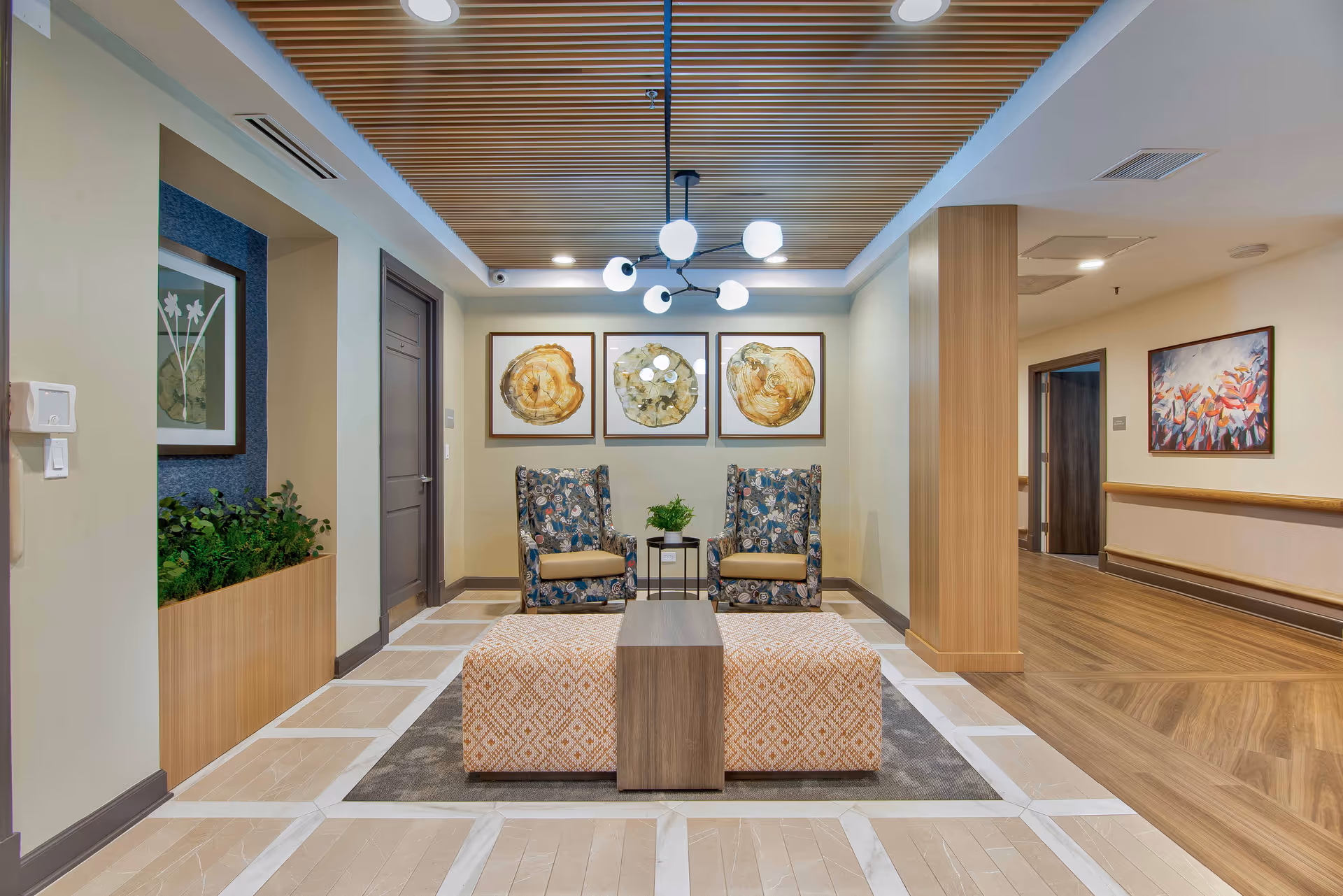 A modern and well-lit seating area in a senior living facility featuring two patterned armchairs with beige cushions, a small table with a plant between them, and two patterned ottomans with a wooden divider in the center. The floor has a light wood and tile design, and the ceiling has wooden slats with a contemporary chandelier. The walls are decorated with framed abstract artwork and there is a hallway visible to the right.