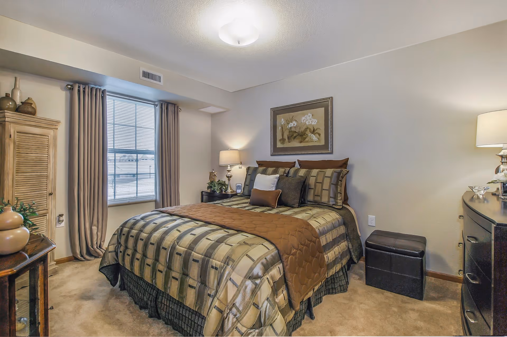 A cozy bedroom with a neatly made bed featuring a patterned bedspread and multiple pillows. The room has beige walls, a window with closed curtains, a wooden cabinet, a nightstand with a lamp and plant, a dresser with a lamp, and a small ottoman.