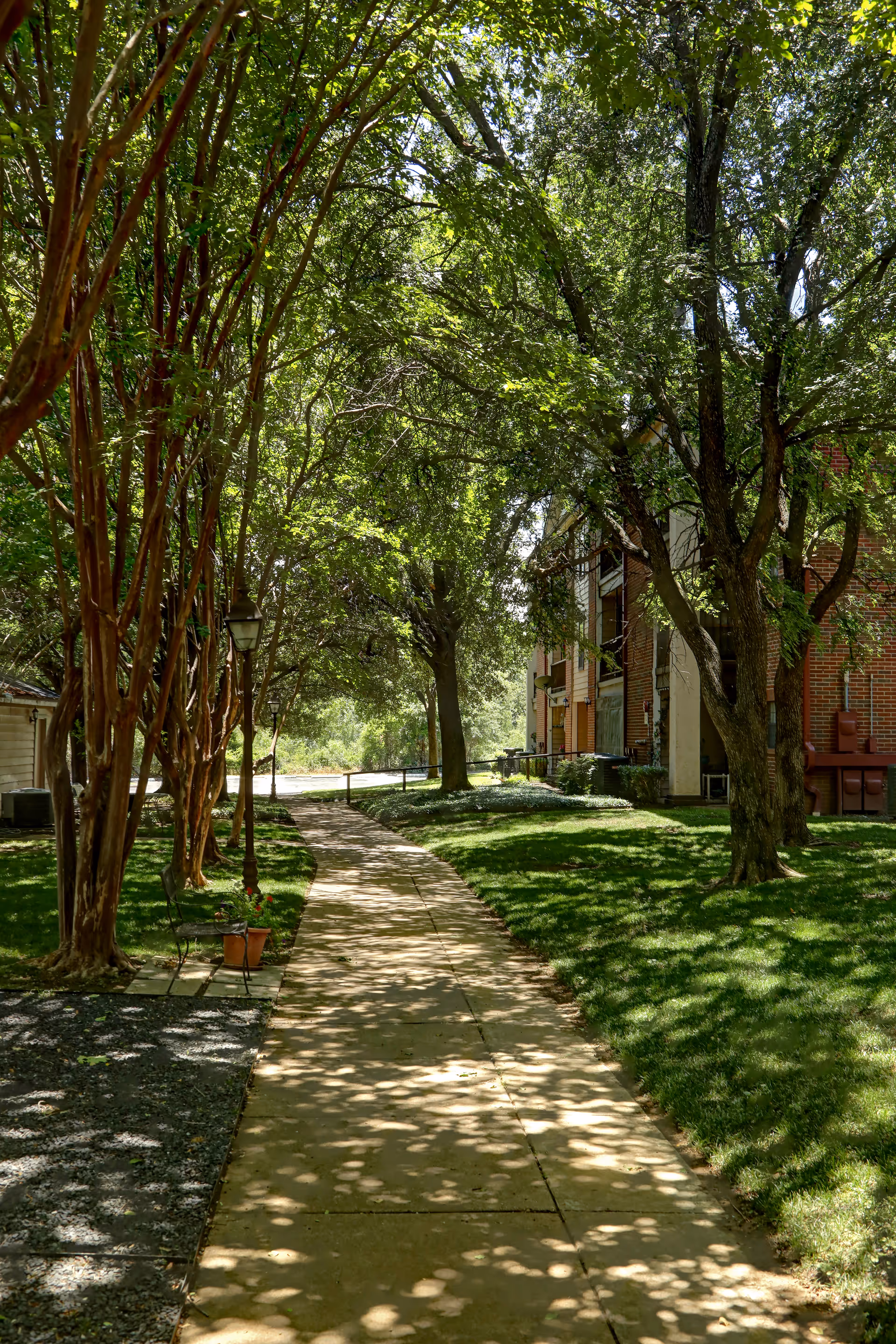 Shaded sidewalk lined with trees leading past a brick residential building and lawn.