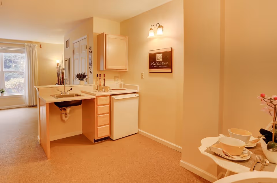 Warm-lit studio kitchenette with a sink, mini-fridge, cabinets and a small table set adjacent to a living area with a window.