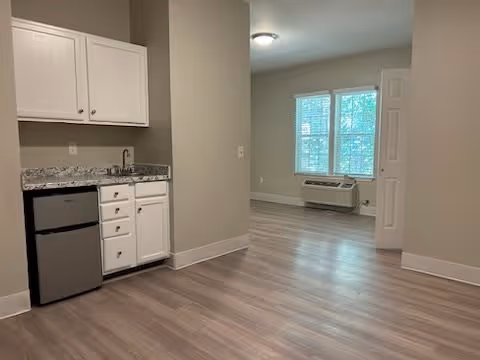 Interior view of a senior living facility room with light wood flooring, a small kitchenette with white cabinets and a mini fridge, and a window air conditioning unit beneath two windows in the adjacent room.