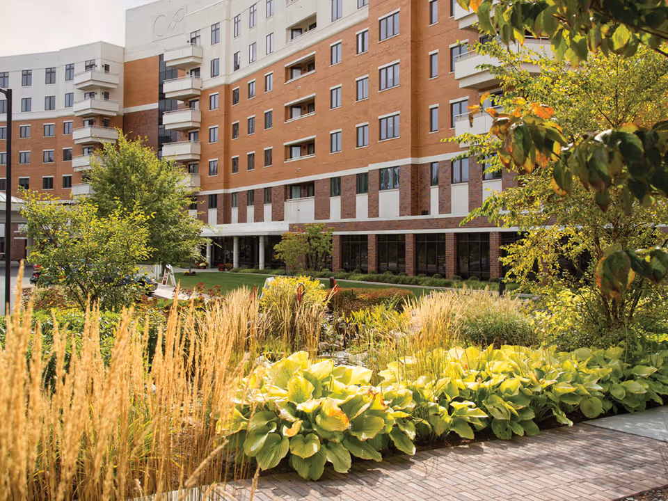 View of the Whitney Center building exterior with multiple floors and balconies, surrounded by a landscaped garden featuring various green plants, tall grasses, and trees under a clear sky.