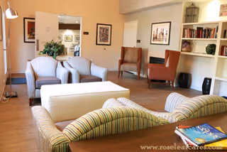 A cozy living room area with striped sofas and armchairs arranged around a white ottoman. The room features wooden flooring, built-in white bookshelves with books and decorative items, framed artwork on the walls, and a floor lamp near the window. There is a doorway leading to another room in the background.