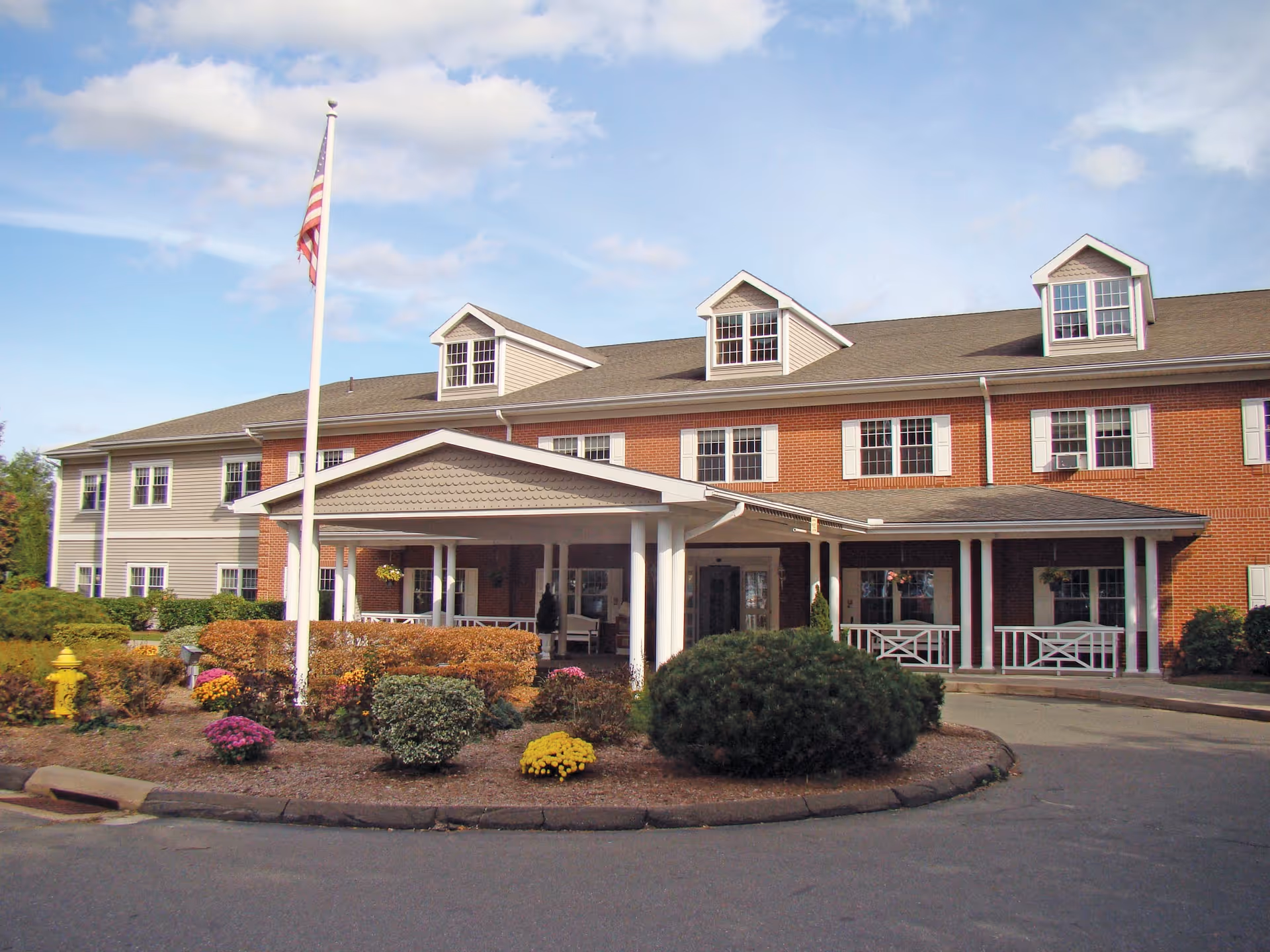 Exterior view of a two-story senior living facility building with a covered entrance, multiple windows, and a flagpole with an American flag in front. There are landscaped bushes and flowers around the entrance area under a partly cloudy sky.