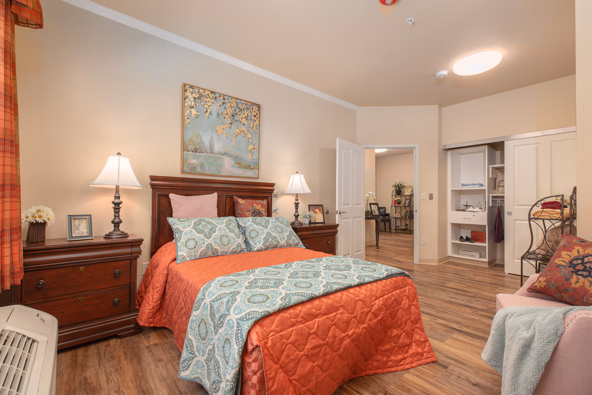 A cozy bedroom in Cedarbrook Memory Care Community featuring a bed with an orange quilt and patterned blue pillows and blanket. The room has wooden nightstands with lamps and framed photos, a painting above the bed, a closet with sliding doors, a pink armchair with a colorful pillow and a light blue throw, and wood flooring. The door to an adjacent room is open, showing a glimpse of another space with a plant and furniture.