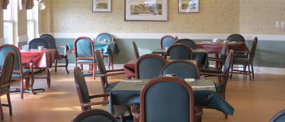 A dining room with multiple tables covered in red and green tablecloths, each surrounded by wooden chairs with green cushions. The room has wooden flooring, patterned wallpaper, framed pictures on the wall, and large windows letting in natural light.