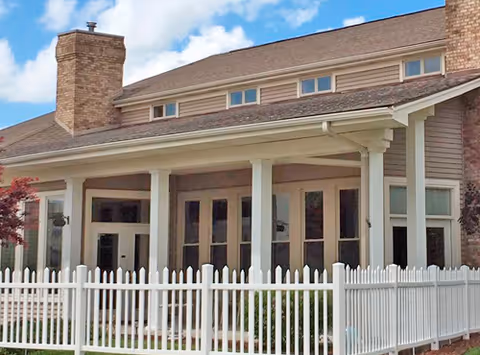 Exterior view of a building with a covered porch supported by white columns, large windows, a brick chimney, and a white picket fence in front. The sky is partly cloudy.