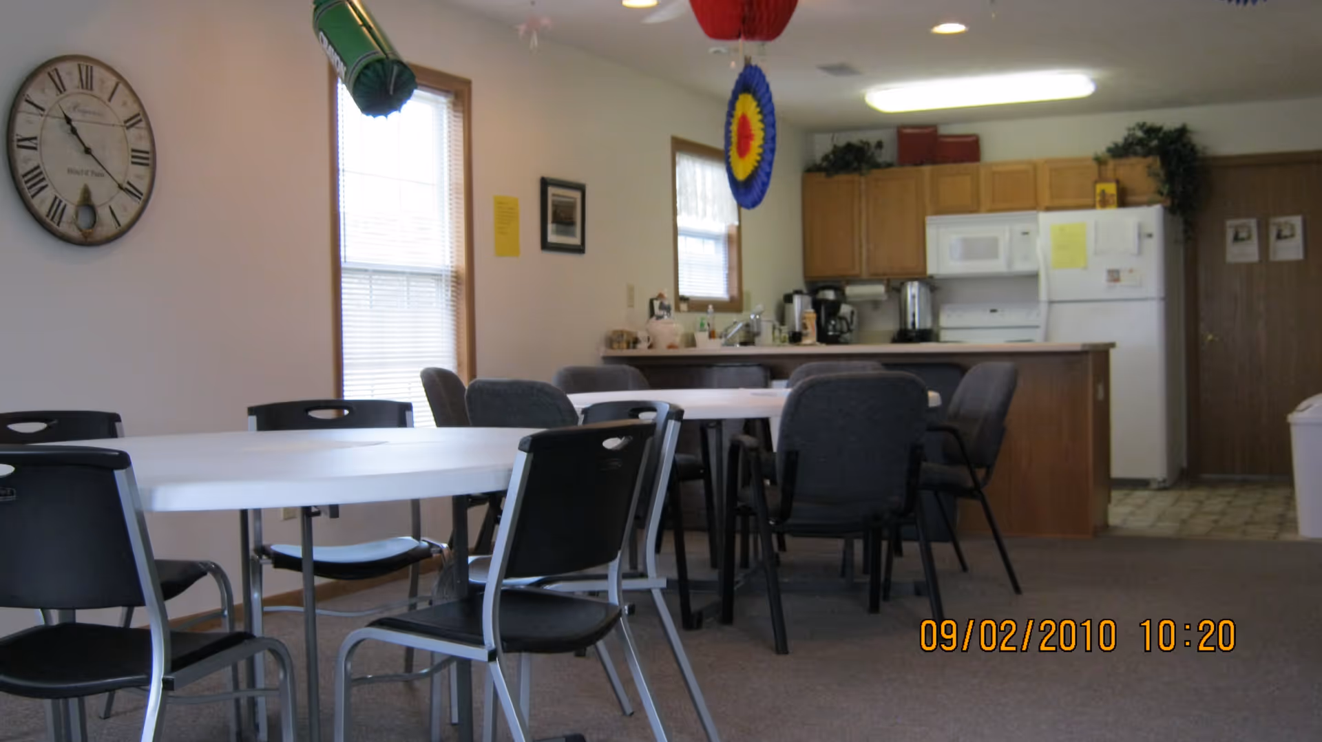 Communal dining area with tables and chairs facing a small kitchen and refrigerator.