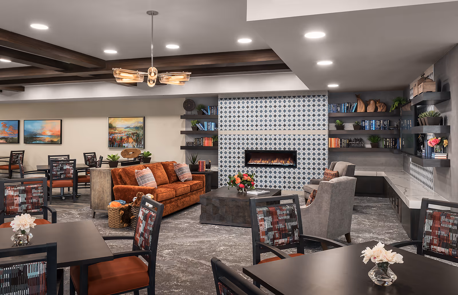 A communal senior living lounge and dining area with tables and chairs, a rust-colored sofa, armchairs, a patterned tile fireplace, and bookshelves.