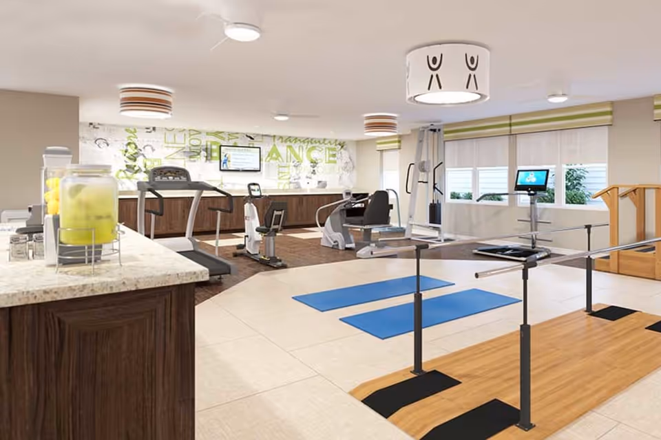 A bright and spacious fitness room in a senior living facility featuring exercise equipment including treadmills, stationary bikes, weight machines, parallel bars for walking support, and two blue exercise mats on the floor. There is a countertop with a beverage dispenser filled with lemon water and cups. The room has large windows with blinds, modern ceiling lights, and a wall decorated with motivational words.