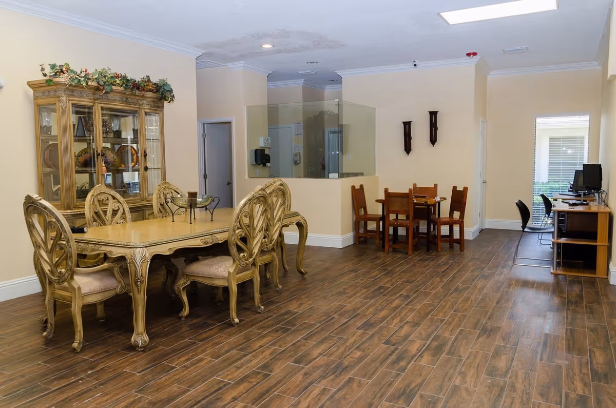 Interior view of a room in an adult daycare facility featuring a large ornate wooden dining table with six matching chairs on the left, a wooden china cabinet with decorative plates and greenery on top, a smaller wooden table with four chairs in the middle, and a computer desk with two chairs near a window on the right. The room has wood-look tile flooring, beige walls, and recessed lighting.