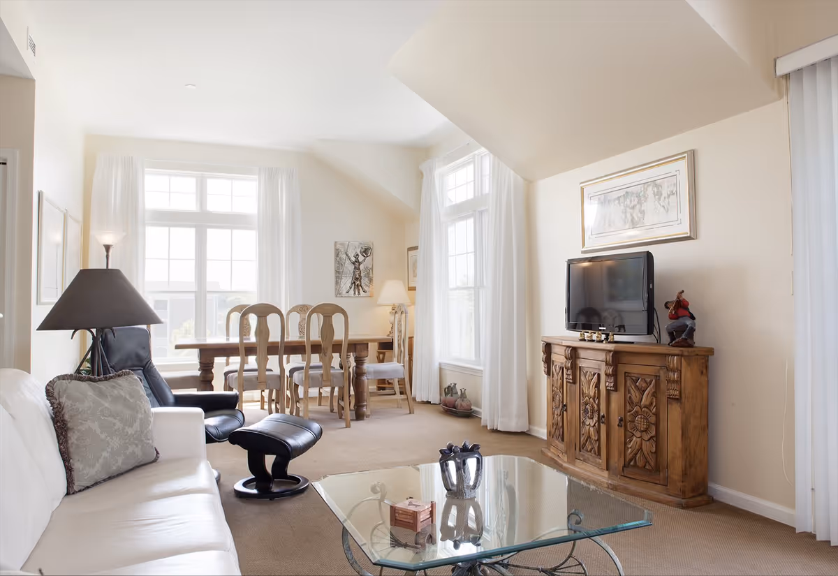 A bright and airy living room and dining area with large windows covered by white curtains. The living room features a white sofa with a decorative pillow, a black leather recliner with matching footrest, a glass coffee table with decorative items, and a wooden cabinet with intricate carvings holding a flat-screen TV. The dining area has a wooden table with six chairs and is illuminated by a floor lamp and natural light from the windows.