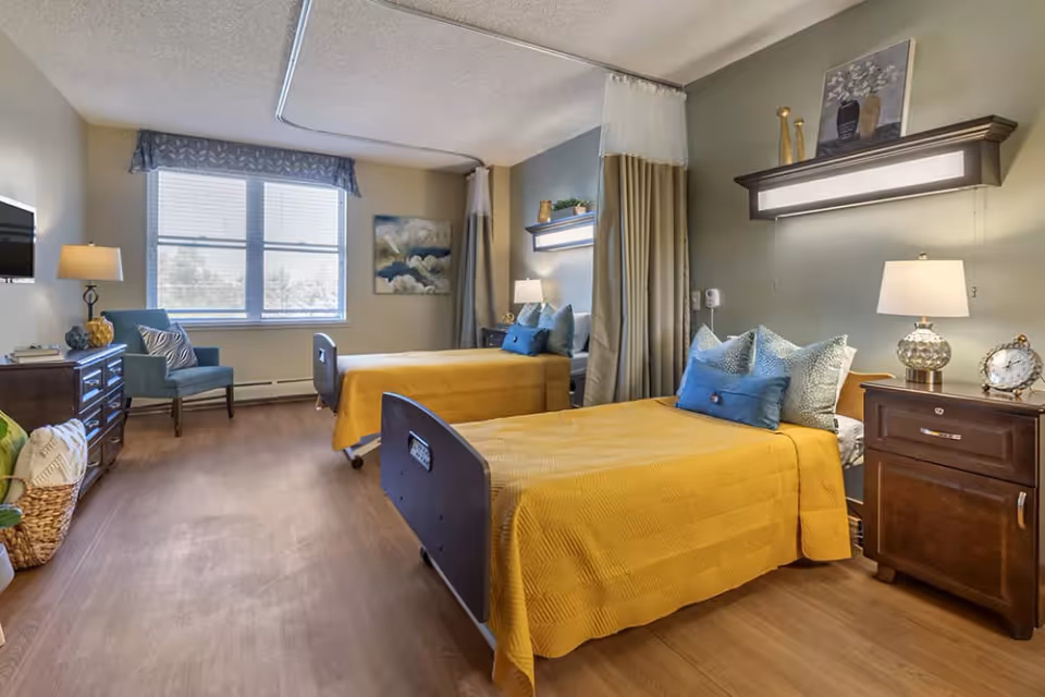 A well-lit room with two single beds covered in yellow bedspreads and blue pillows. Each bed has a bedside table with a lamp and decorative items. There is a curtain divider between the beds. The room has a wooden floor, a window with blinds, a blue armchair, a dresser with a lamp, and wall art.