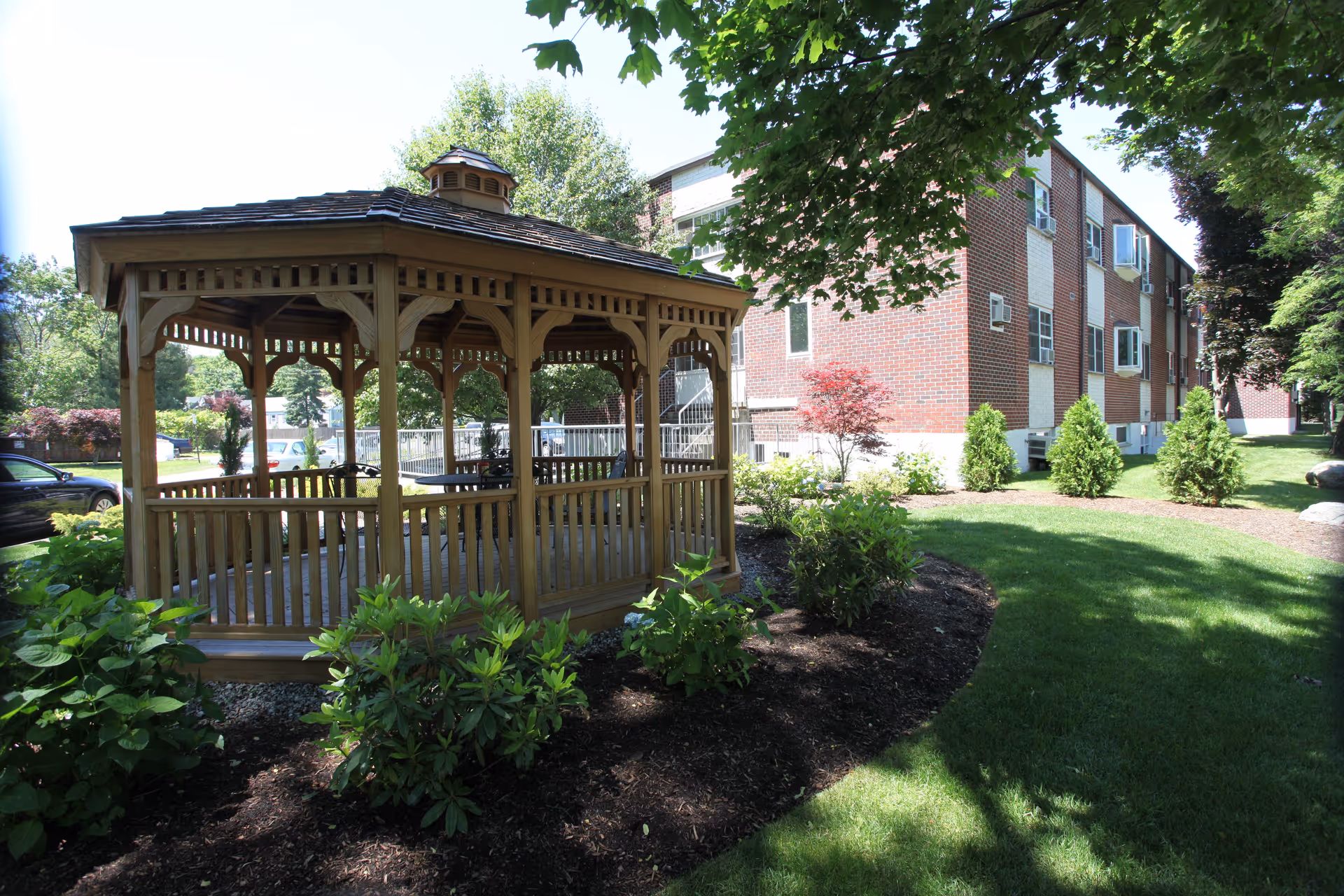 A wooden gazebo surrounded by green shrubs and trees in a landscaped garden area next to a multi-story brick building with several windows and air conditioning units.