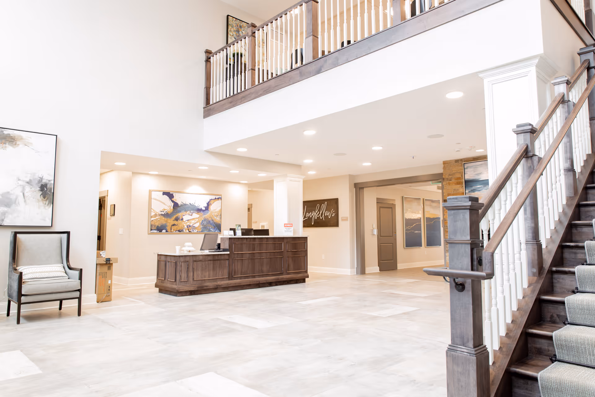 Bright and spacious senior living facility lobby with a wooden reception desk, a comfortable armchair with a pillow, abstract artwork on the walls, and a staircase with wooden handrails and white balusters leading to an upper floor.