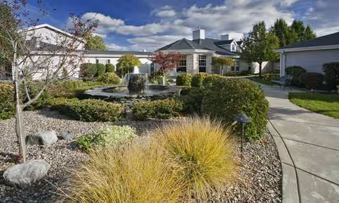 Front view of a senior living campus with a central fountain, curved walkway, and trimmed shrubs.