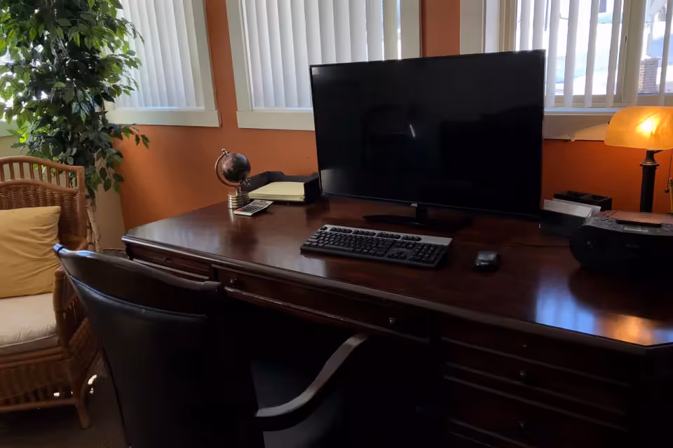 A wooden desk with a computer monitor, keyboard, mouse, small globe, calculator, and a lamp. There is a black office chair in front of the desk and a wicker chair with a yellow cushion to the side. Vertical blinds cover the windows behind the desk, and a green plant is visible in the corner.