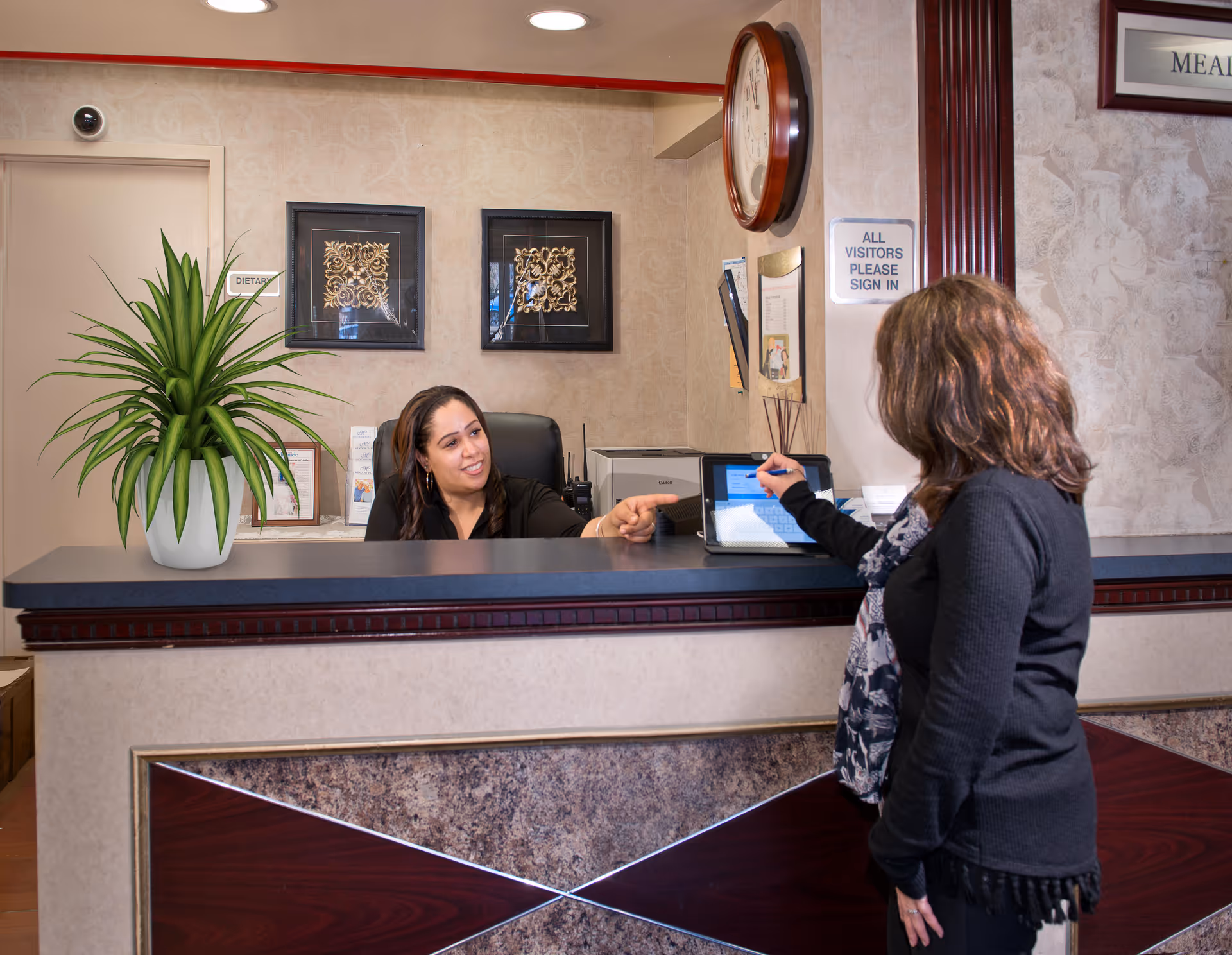 A receptionist sitting behind a front desk is smiling and pointing while a visitor signs in on a digital tablet. The reception area has a potted plant, two framed decorative wall pieces, a clock, and a sign that reads 'ALL VISITORS PLEASE SIGN IN'.