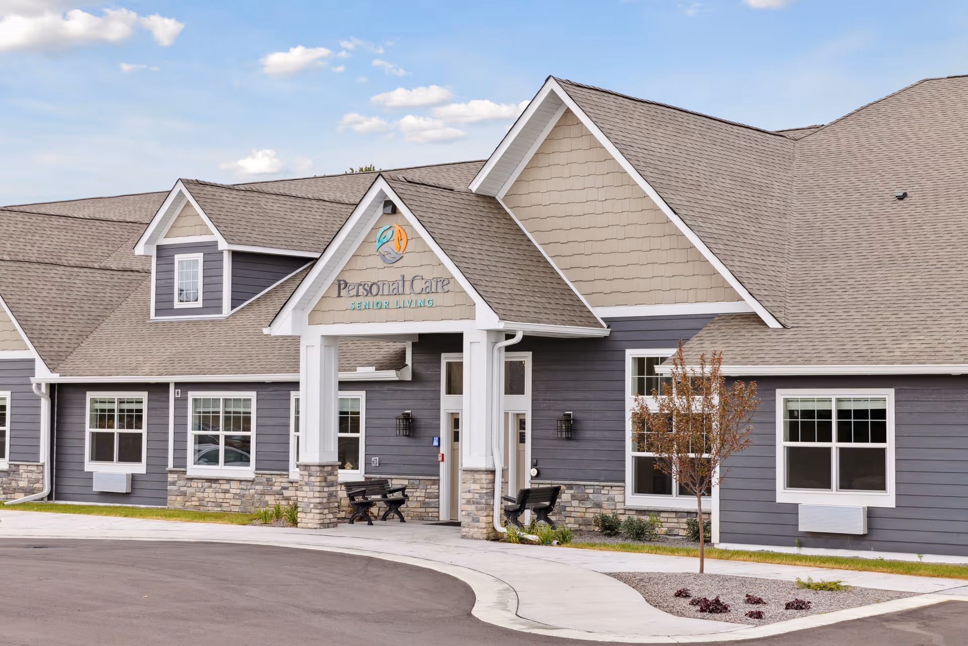Exterior front view of a senior living facility named Personal Care Senior Living - Columbia Heights, featuring a modern building with gray siding, stone accents, multiple windows, a covered entrance with benches, and a small landscaped area with a young tree.